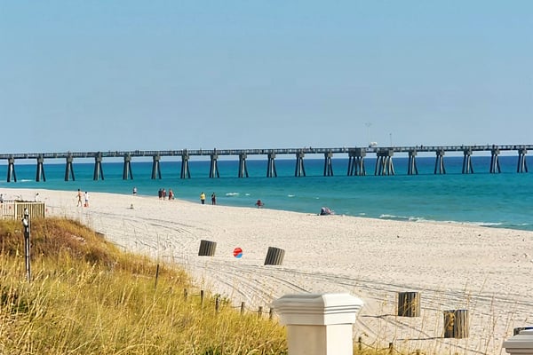 Sandy beach with dune grass, long fishing pier extending into turquoise ocean, and beachgoers in the distance