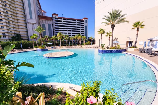 Resort pool with turquoise water, central island, palm trees, and high-rise buildings under clear blue sky