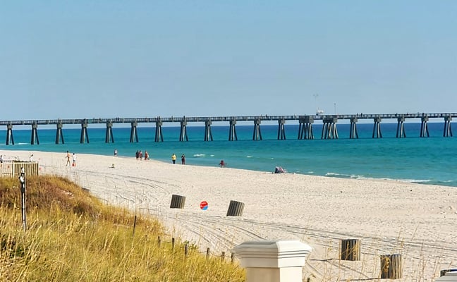 Sandy beach with dune grass, long fishing pier extending into turquoise ocean, and beachgoers in the water